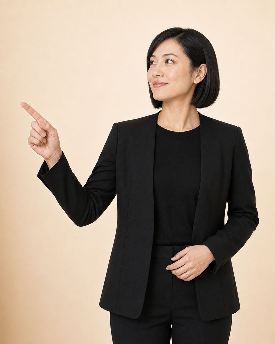 An auditor in a black trouser suit over a black top, with a dark bob, standing against a warm pale wall and pointing into the open space alongside.