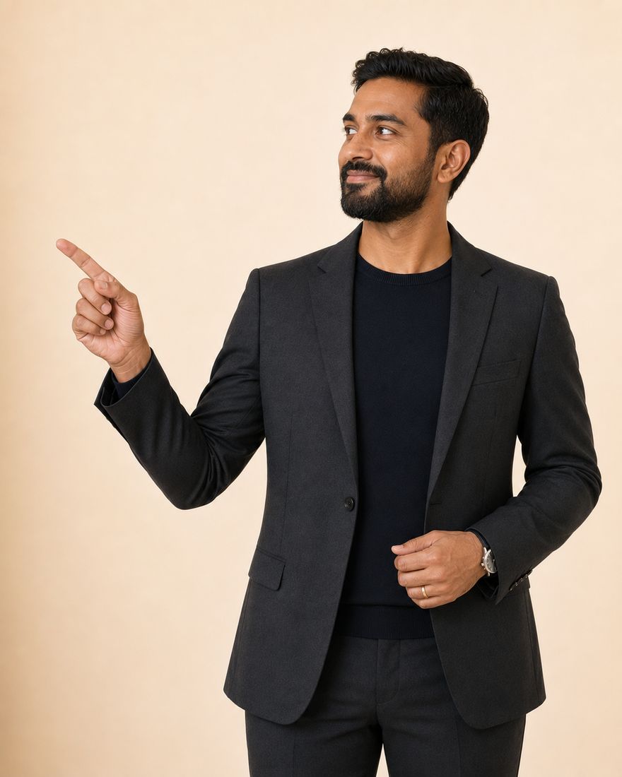 An auditor in a charcoal blazer over a navy crew-neck, with a trimmed beard, standing against a warm pale wall and pointing into the open space alongside.