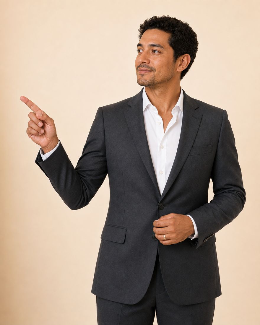 An auditor in a charcoal suit and open-collared white shirt, with dark curly hair, standing against a warm pale wall and pointing into the open space alongside.